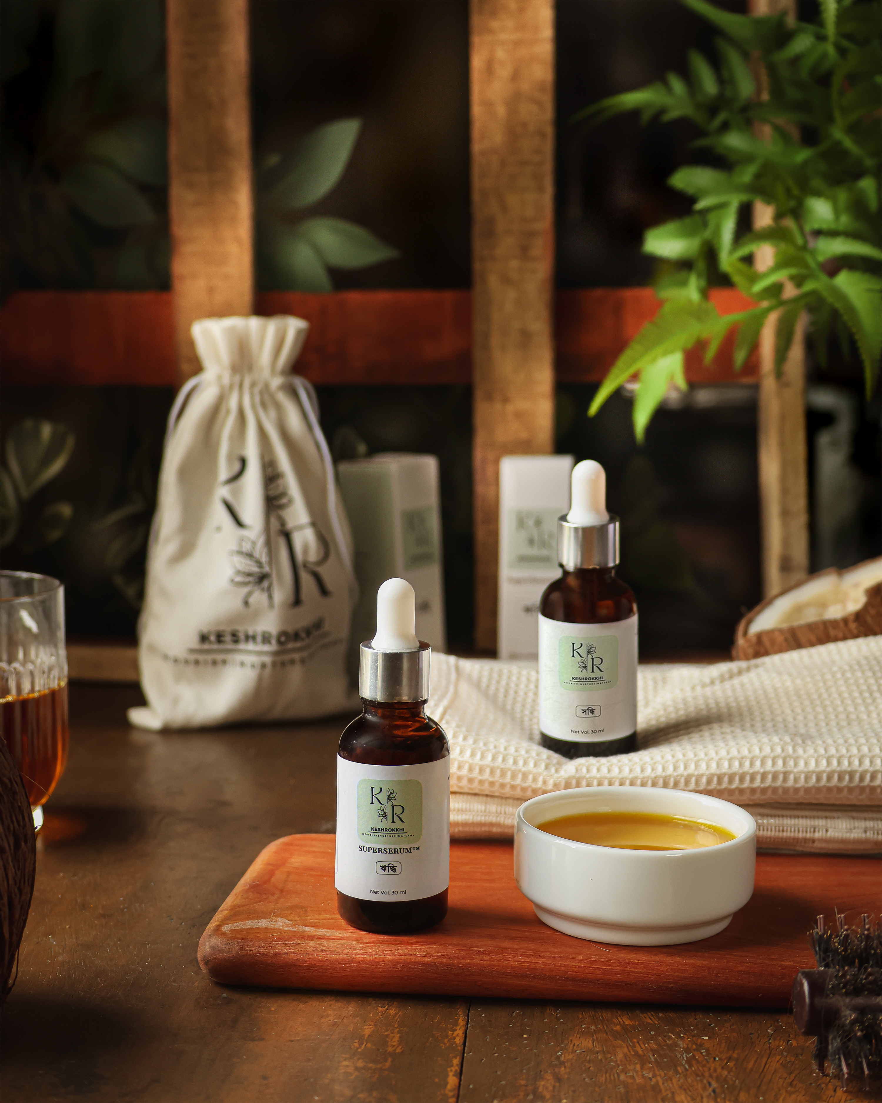 Keshrokkhi hair care serums displayed in a rustic wooden setting with natural oil and pouch packaging- made in Bangladesh for hair fall control, frizz control and shine