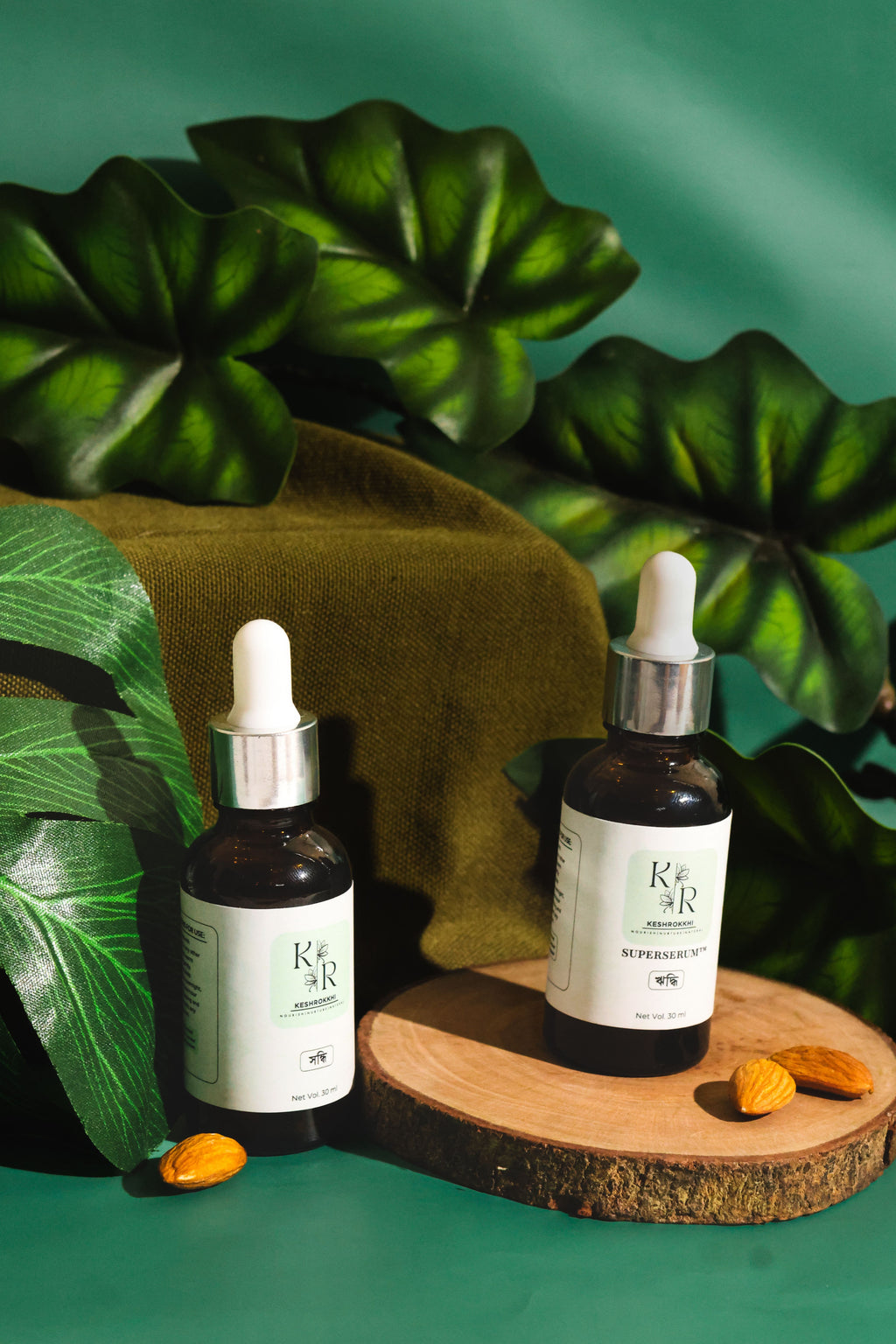 Keshrokkhi Hriddhi and Shondhi hair serums on a wooden slab with almonds and tropical green leaves- organic hair fall and repair oils from Bangladesh