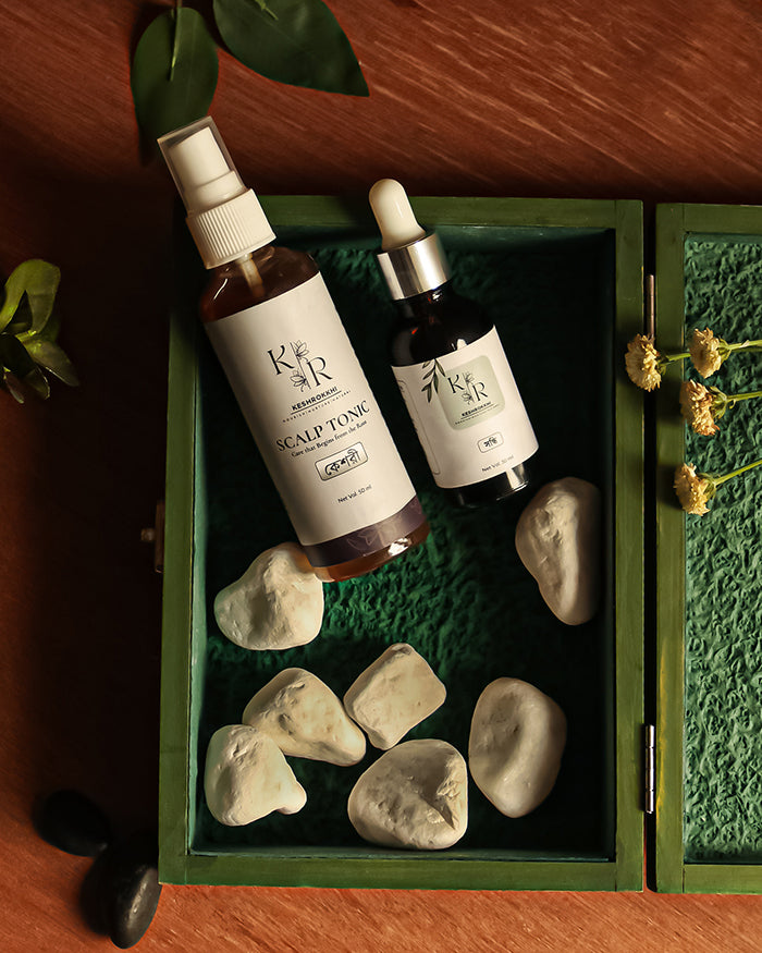 Two bottles of Keshrokkhi Scalp Tonic and Shondhi hair serum on a green tray with stones and plants. Designed to repair and grow hair. 