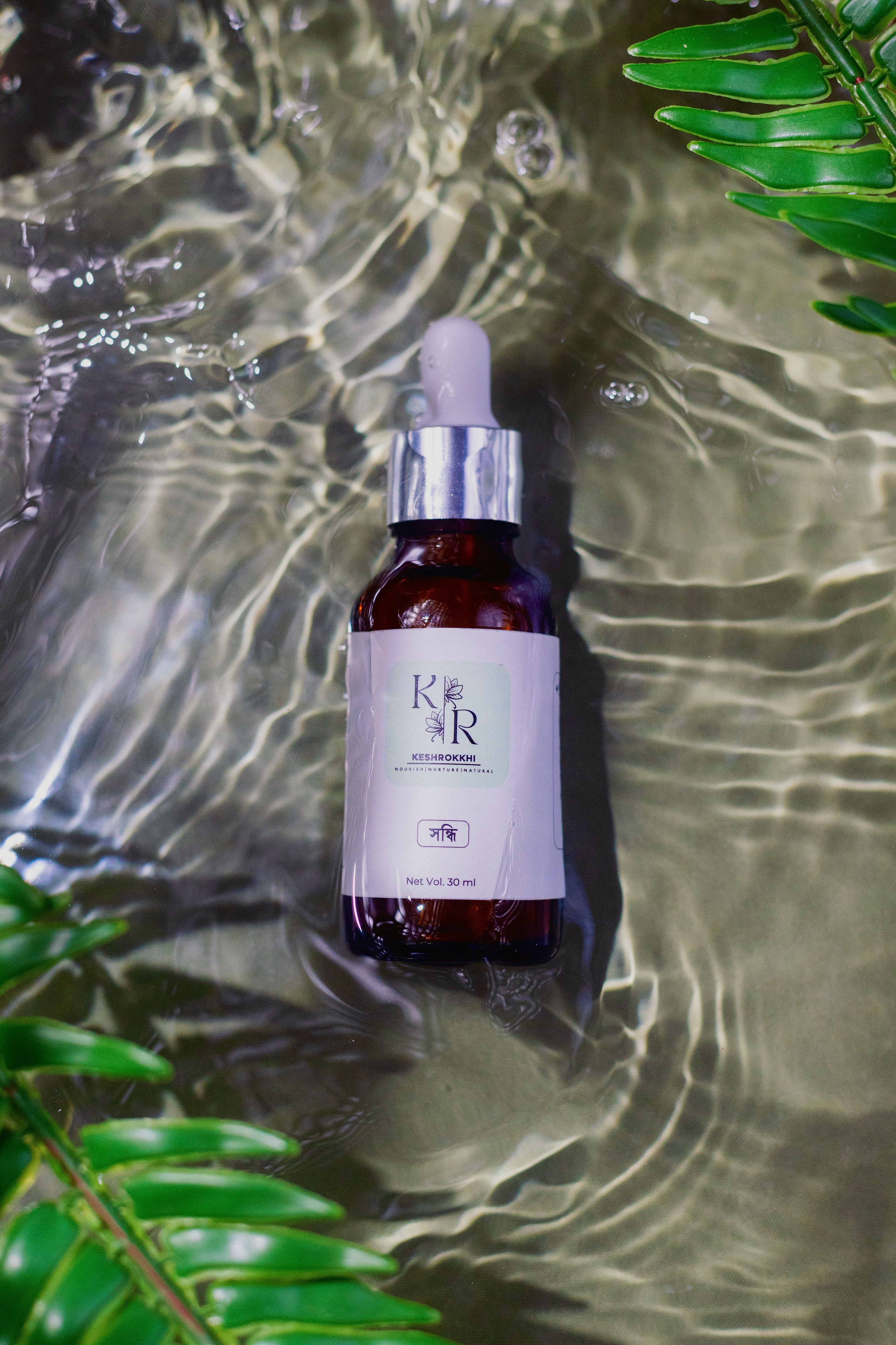 Keshrokkhi Shondhi serum with, repair and breakage solution submerged in rippling water with fresh green leaves- organic hair oil for women and men made in Bangladesh but ingredients sourced from around the world. 
