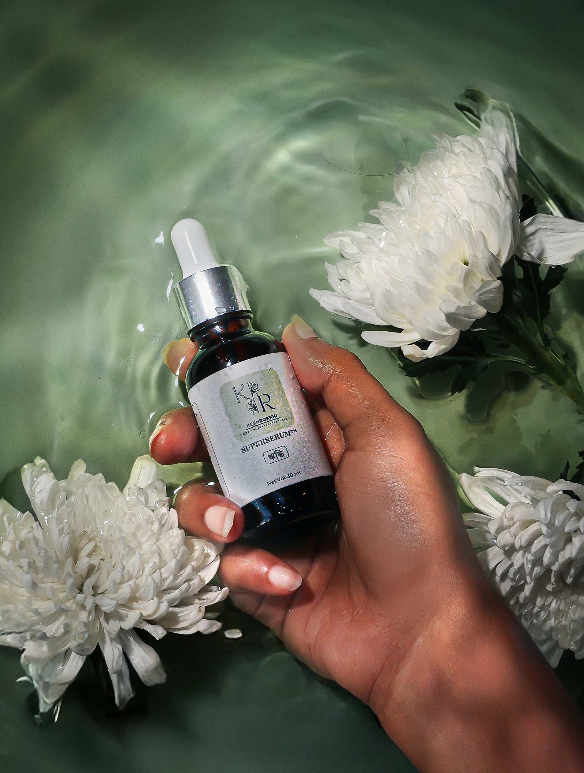 Hand holding a bottle of Hriddhi Hair serum with flowers in water. Best for Hair Fall control, Hair Frizz Control in Bangladesh