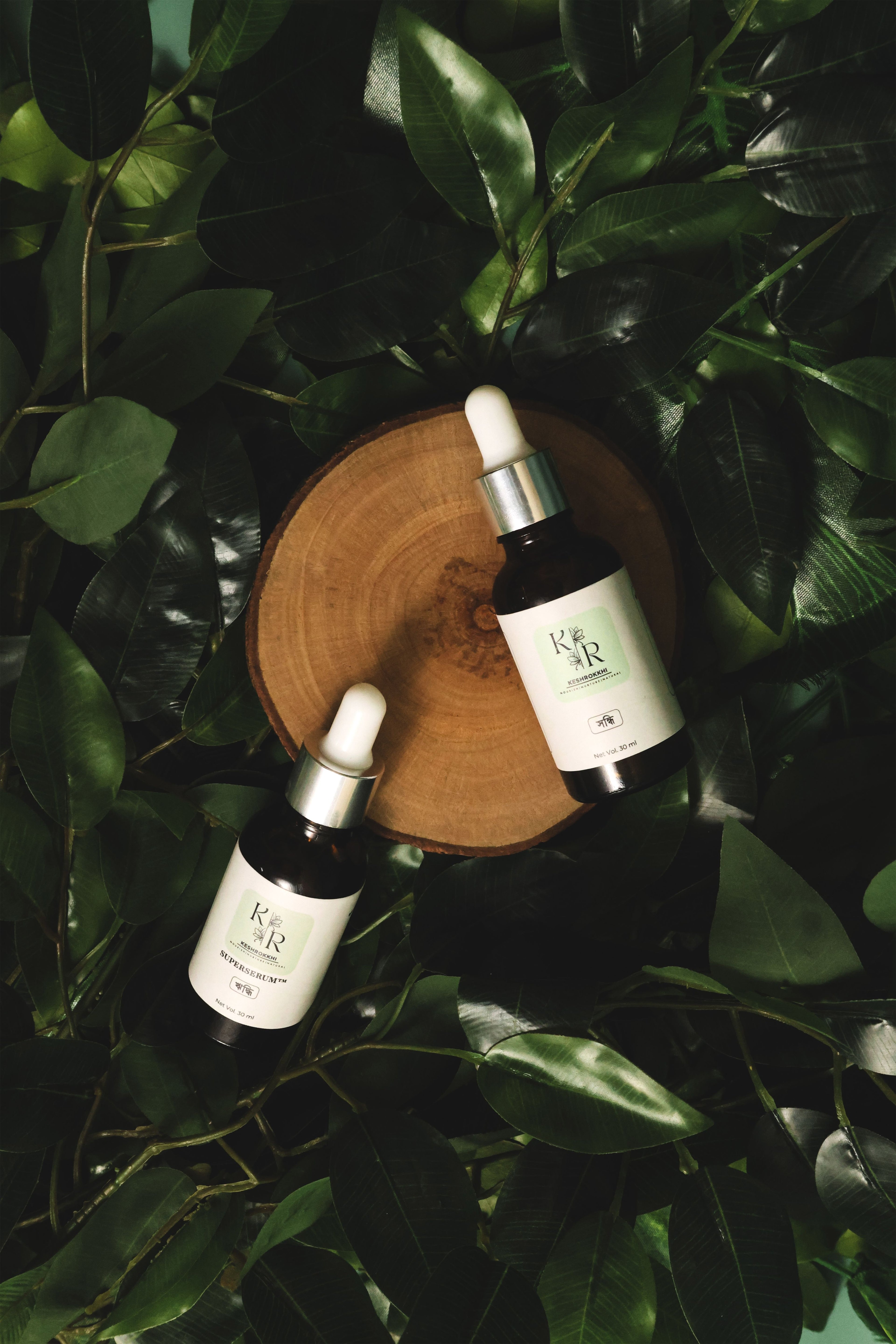 Keshrokkhi hriddhi and shondhi hair serums displayed on a wooden log slice surrounded by lush green leaves- all- natural hair care duo for fall control and repair. 